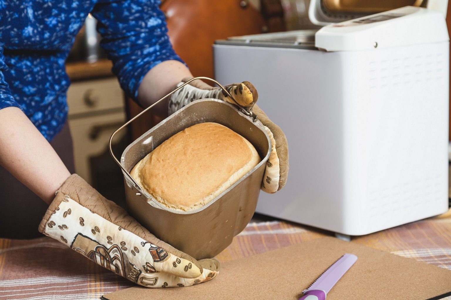 Female hands in kitchen gloves holding metal mold for baking bread. Inside appetizing hot loaf of bread. Behind is an electric bread maker